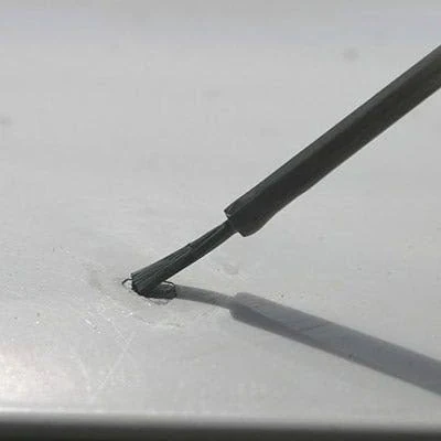 A small applicator brush dabbing touch-up paint onto a rock chip in a light gray painted car panel