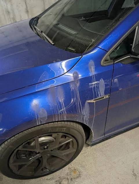 Chalky white mineral drips and a film of dust streaking down the front fender and wheel of a blue car parked beside a bare concrete wall in a garage