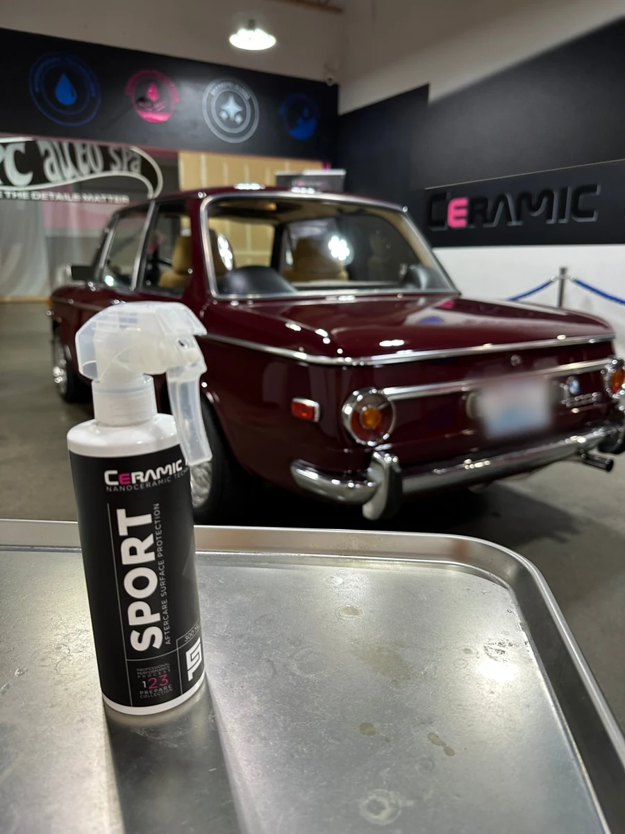 A white spray bottle of Ceramic Pro Sport aftercare product standing on a stainless steel table, with a maroon classic BMW parked behind it inside a detailing shop