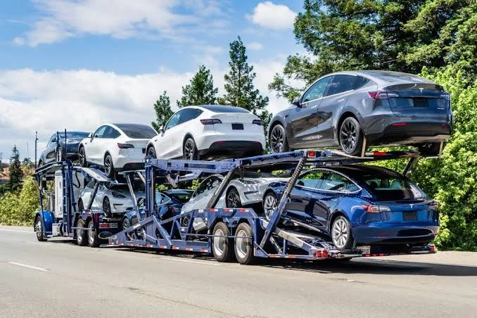 A multi-level car carrier truck loaded with new sedans and SUVs driving along a highway lined with trees