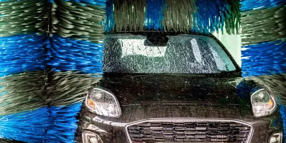 A dark car moving through a tunnel car wash, surrounded by large blue and green brushes pressed against the paint