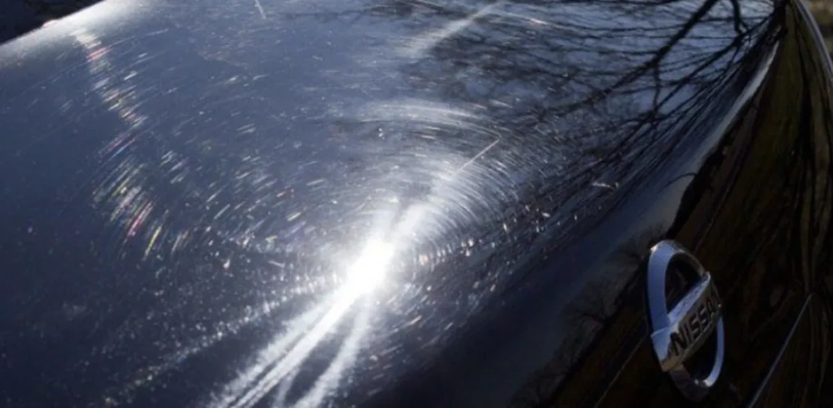 Close-up of glossy black paint covered in fine circular swirl marks and a spiderweb pattern around a bright reflection of the sun, with a chrome emblem at the lower right